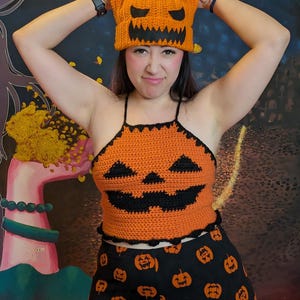 A person smiling playfully while holding the orange pumpkin beanie above their head in full outfit.