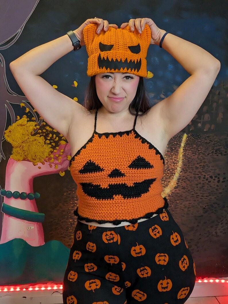 A person smiling playfully while holding the orange pumpkin beanie above their head in full outfit.
