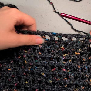 Close-up of hands working on dark speckled yarn with an open mesh crochet stitch using a red crochet hook.
