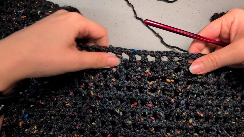 Close-up of hands working on dark speckled yarn with an open mesh crochet stitch using a red crochet hook.