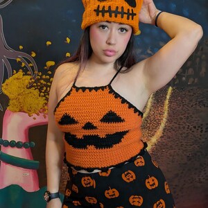 A person posing with one hand behind their head wearing the orange crochet pumpkin hat, top, and patterned pants.