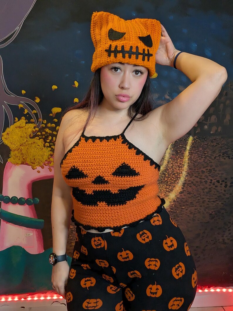 A person posing with one hand behind their head wearing the orange crochet pumpkin hat, top, and patterned pants.