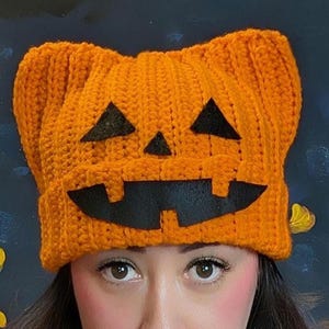 Close-up of a person wearing the orange crochet pumpkin cat beanie against a dark background.