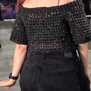 Back view of a person wearing a black strap under an off-the-shoulder dark speckled crochet top and black jeans.