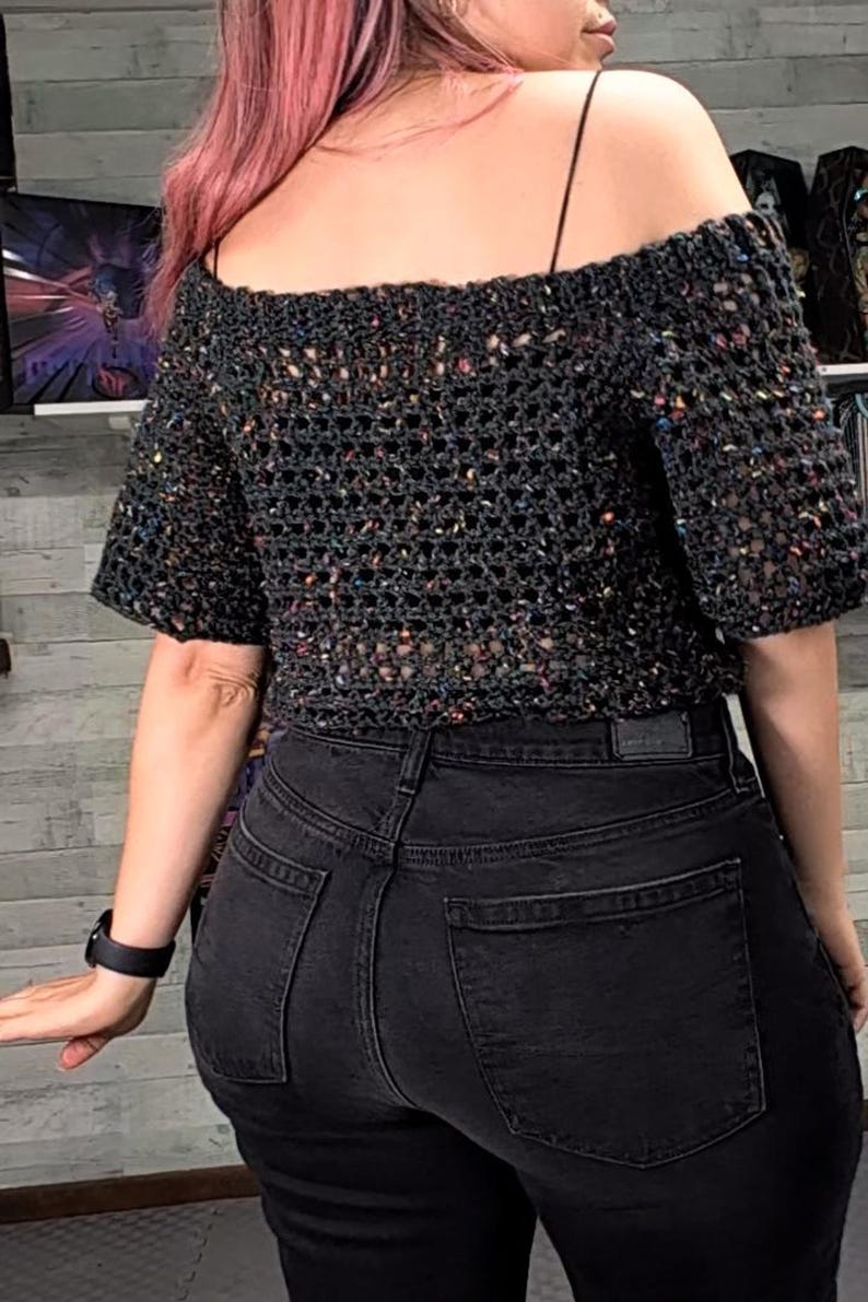 Back view of a person wearing a black strap under an off-the-shoulder dark speckled crochet top and black jeans.