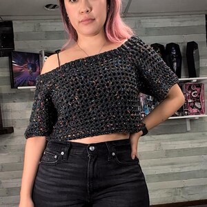 A person with pink hair stands facing forward wearing a short-sleeved dark speckled mesh crochet top and black jeans against a wood-paneled wall.