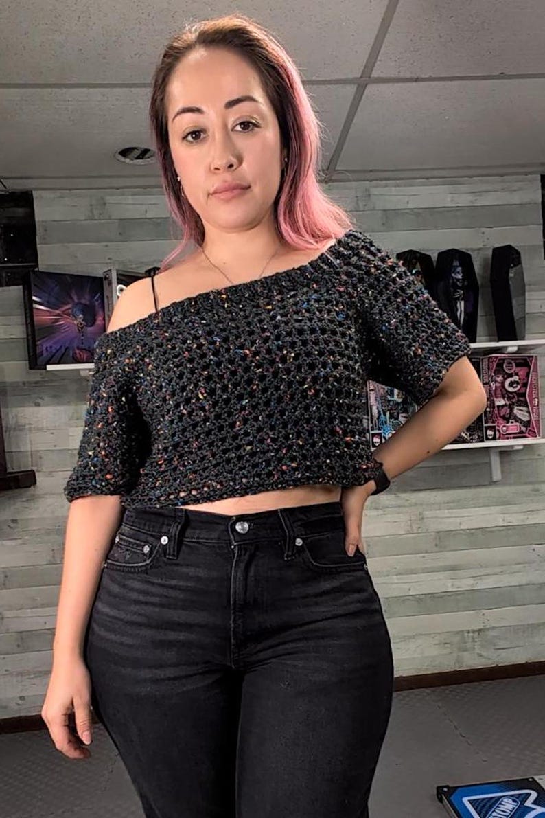 A person with pink hair stands facing forward wearing a short-sleeved dark speckled mesh crochet top and black jeans against a wood-paneled wall.