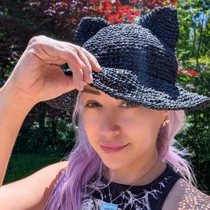 May include: A black crocheted bucket hat featuring two pointed cat-like ears on the top. The hat is constructed from a textured, woven material with a wide, slightly flared brim and a consistent dark black color throughout the entire design.