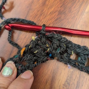 Close-up on a wooden table of dark speckled yarn being crocheted with a red hook and held by a hand with light green nail polish.