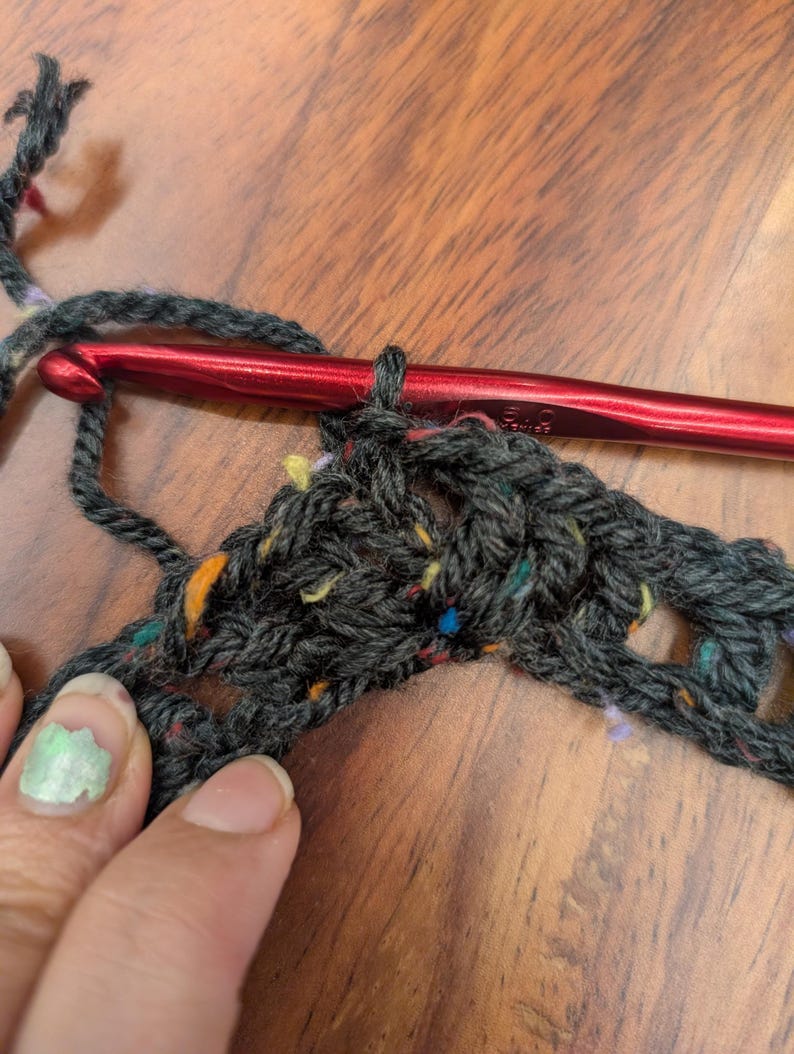 Close-up on a wooden table of dark speckled yarn being crocheted with a red hook and held by a hand with light green nail polish.