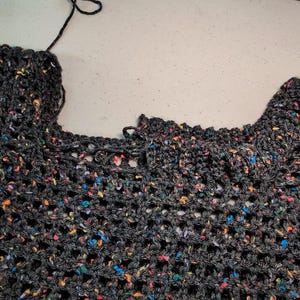 A piece of dark speckled mesh crochet fabric lies flat on a light-colored surface with a hand visible on the left.