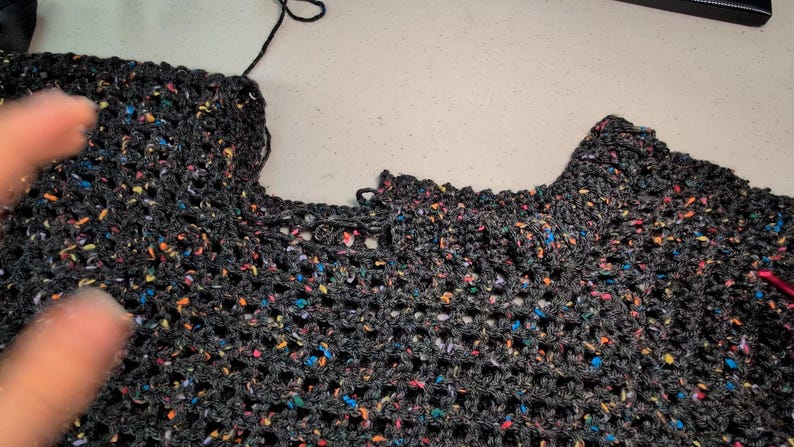 A piece of dark speckled mesh crochet fabric lies flat on a light-colored surface with a hand visible on the left.