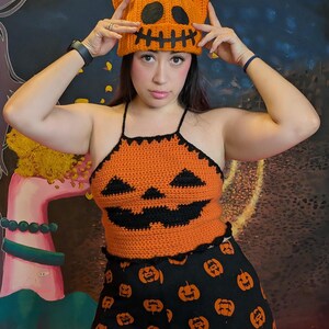 A person holding up the orange crochet pumpkin beanie while wearing a matching pumpkin halter top and leggings.
