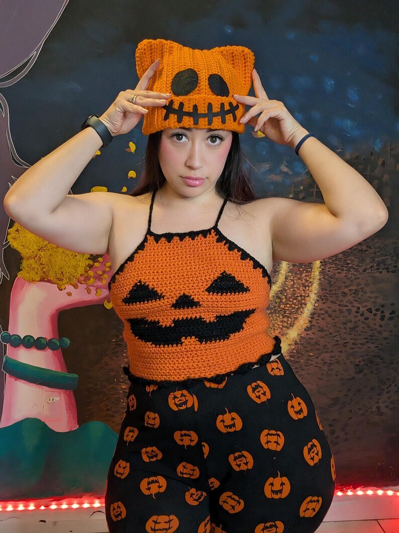 A person holding up the orange crochet pumpkin beanie while wearing a matching pumpkin halter top and leggings.
