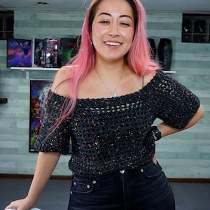 A smiling person with pink hair poses with one hand on their hip, wearing an off-the-shoulder dark speckled crochet top.