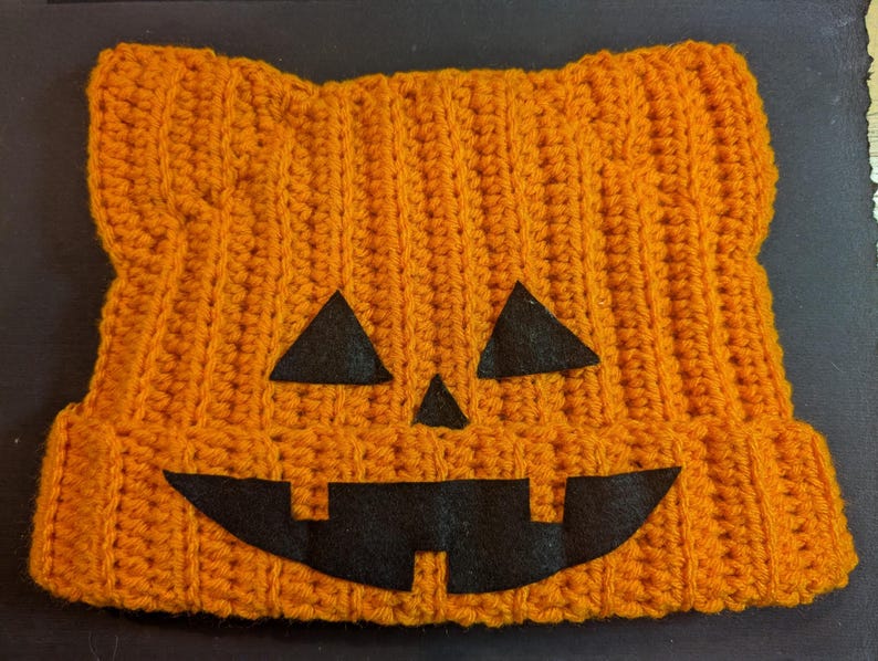 Flat lay of the orange ribbed crochet cat beanie featuring black felt jack-o-lantern eyes, nose, and mouth.