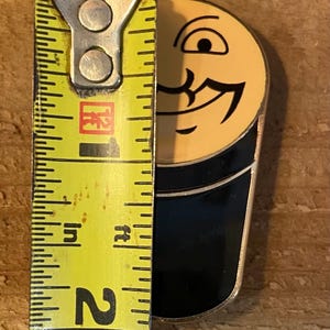 Puede incluir: Un pin de esmalte negro y amarillo con forma de cara de dibujos animados con un solo ojo y una amplia sonrisa. El pin mide aproximadamente 5 cm de alto.