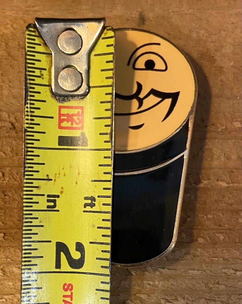 Puede incluir: Un pin de esmalte negro y amarillo con forma de cara de dibujos animados con un solo ojo y una amplia sonrisa. El pin mide aproximadamente 5 cm de alto.
