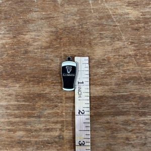 May include: A black and white Guinness pint charm with a white border. The charm is approximately 1 inch in height. The Guinness logo is visible on the front of the charm. The charm is on a wooden surface.