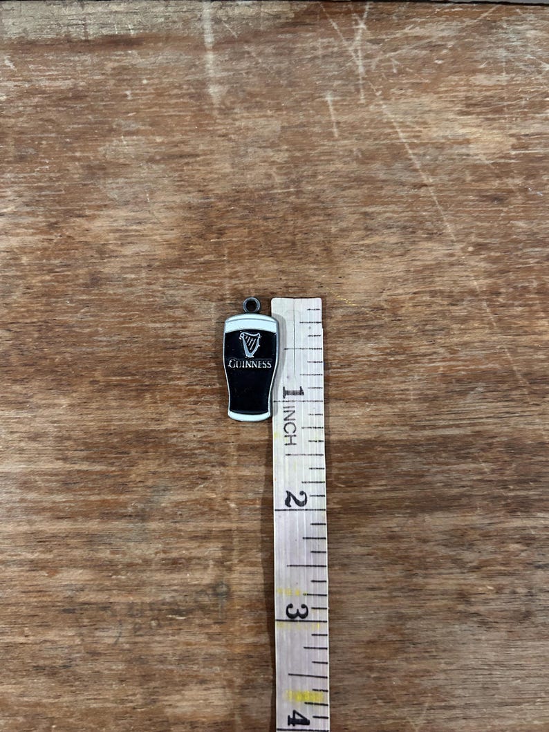 May include: A black and white Guinness pint charm with a white border. The charm is approximately 1 inch in height. The Guinness logo is visible on the front of the charm. The charm is on a wooden surface.