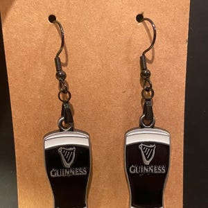 May include: A pair of black and white Guinness earrings. Each earring features a black enamel pint glass design with the Guinness logo and a white top. The earrings hang from black metal hooks.