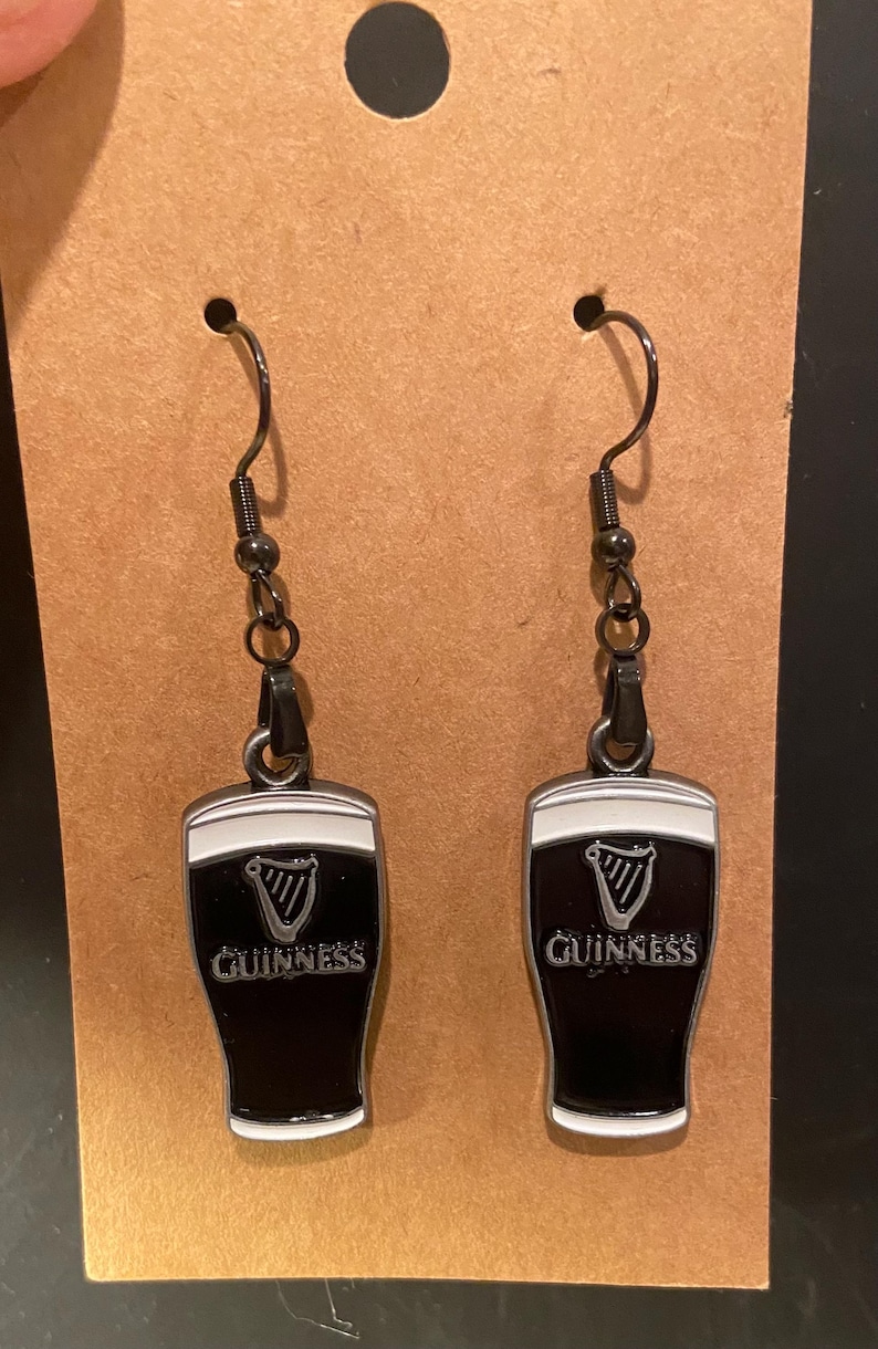 May include: A pair of black and white Guinness earrings. Each earring features a black enamel pint glass design with the Guinness logo and a white top. The earrings hang from black metal hooks.