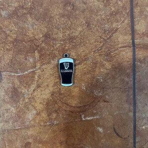 May include: A small, black and white Guinness pint charm with a silver-colored loop for attaching to a bracelet or necklace. The Guinness logo is prominently displayed on the front of the charm. The charm is set against a brown, textured background.