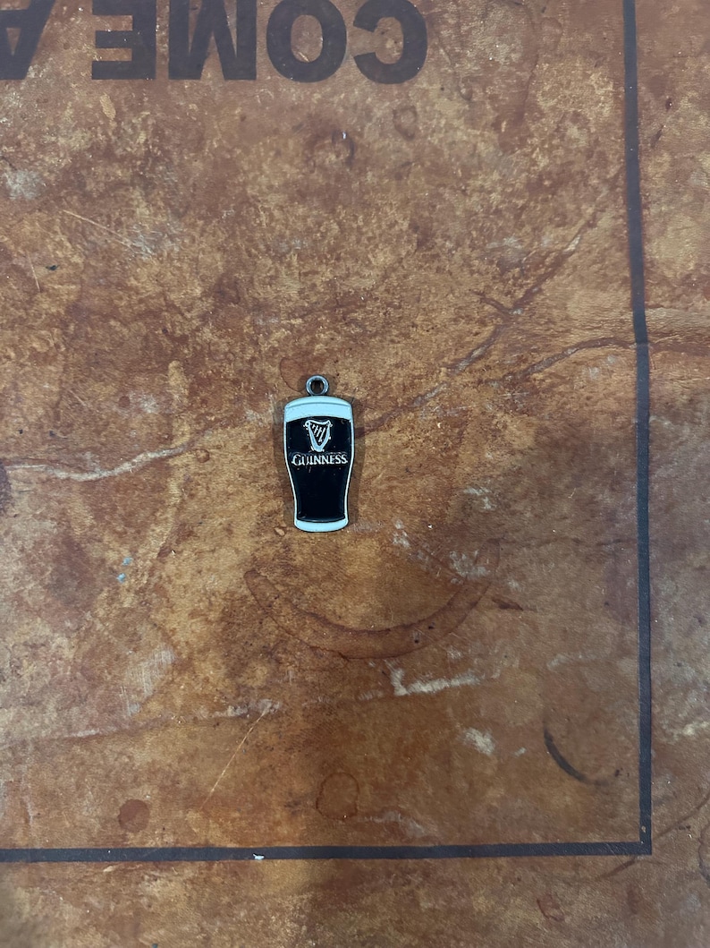 May include: A small, black and white Guinness pint charm with a silver-colored loop for attaching to a bracelet or necklace. The Guinness logo is prominently displayed on the front of the charm. The charm is set against a brown, textured background.