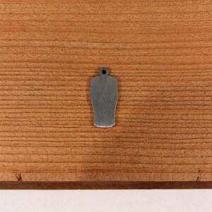 May include: A small, silver-colored charm in the shape of a beer glass, with a hole at the top for attaching to a chain or bracelet. The charm is set against a wooden background with visible grain.