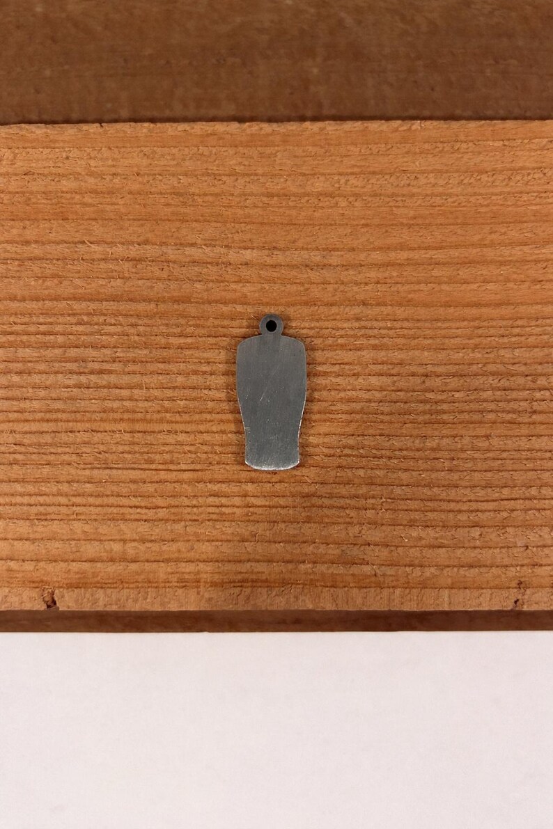 May include: A small, silver-colored charm in the shape of a beer glass, with a hole at the top for attaching to a chain or bracelet. The charm is set against a wooden background with visible grain.
