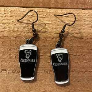 May include: A pair of black drop earrings featuring a Guinness pint design. The earrings have black hooks and the pint glasses are black with a white rim and the Guinness logo.