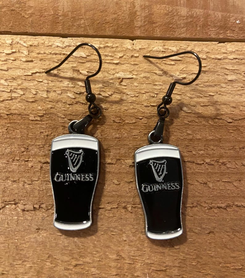 May include: A pair of black drop earrings featuring a Guinness pint design. The earrings have black hooks and the pint glasses are black with a white rim and the Guinness logo.