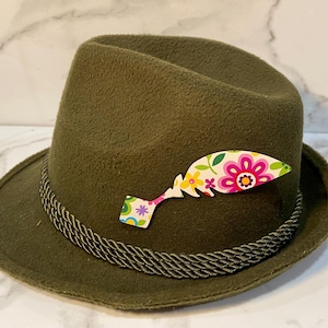 May include: Olive green felt hat with a decorative floral pin. The hat features a braided rope band around the base of the crown. The pin is shaped like a feather and has a colorful floral design with pink, yellow, and white flowers.