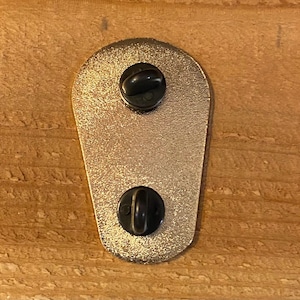 Puede incluir: La parte posterior de un pin de esmalte de color dorado con dos cierres de metal negro.