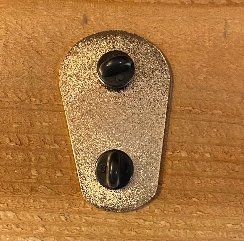 Puede incluir: La parte posterior de un pin de esmalte de color dorado con dos cierres de metal negro.