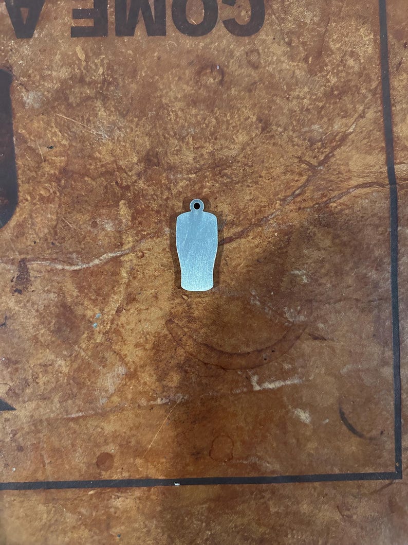 May include: A small, silver-colored charm in the shape of a beer glass. The charm has a small hole at the top for attaching to a necklace or bracelet. The charm is made of a shiny metal and has a smooth surface.