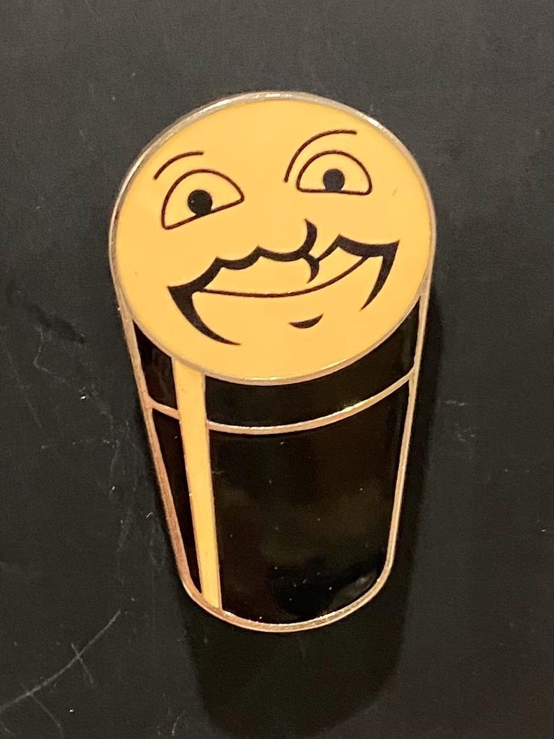 Puede incluir: Un pin de esmalte negro y amarillo de un vaso de cerveza sonriente con bigote.