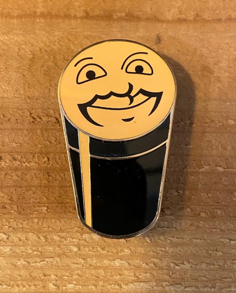 Puede incluir: Un pin de color dorado con forma de vaso de cerveza con una cara sonriente en la parte superior. El vaso de cerveza es negro con un borde dorado.