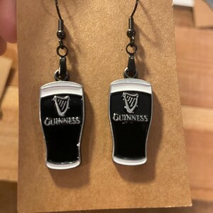May include: A pair of black and white Guinness beer glass earrings. The earrings have a black background with the Guinness logo and text in white. The earrings are hanging from black metal hooks.