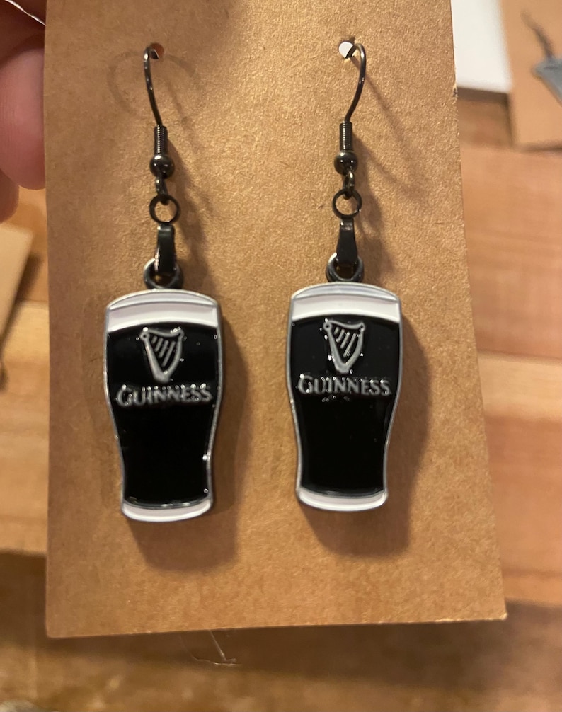 May include: A pair of black and white Guinness beer glass earrings. The earrings have a black background with the Guinness logo and text in white. The earrings are hanging from black metal hooks.