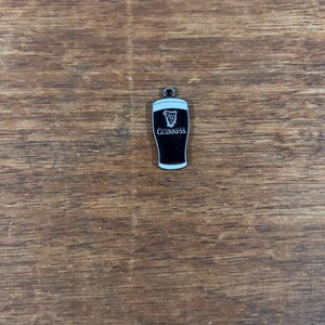 May include: A small, black and white Guinness pint charm. The charm features the Guinness logo and the word "Guinness" in white lettering. The charm has a small loop at the top for attaching to a necklace or bracelet.