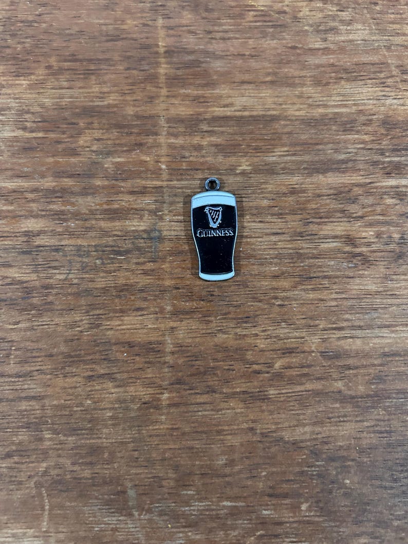 May include: A small, black and white Guinness pint charm. The charm features the Guinness logo and the word "Guinness" in white lettering. The charm has a small loop at the top for attaching to a necklace or bracelet.