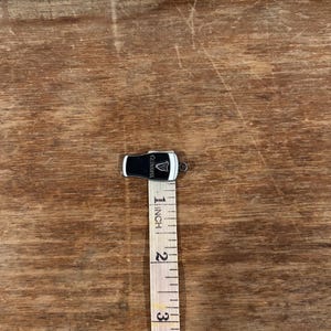 May include: A small, black and white Guinness pint glass charm with the Guinness logo. The charm is approximately 1 inch in length. The charm is on a wooden surface with a measuring tape.