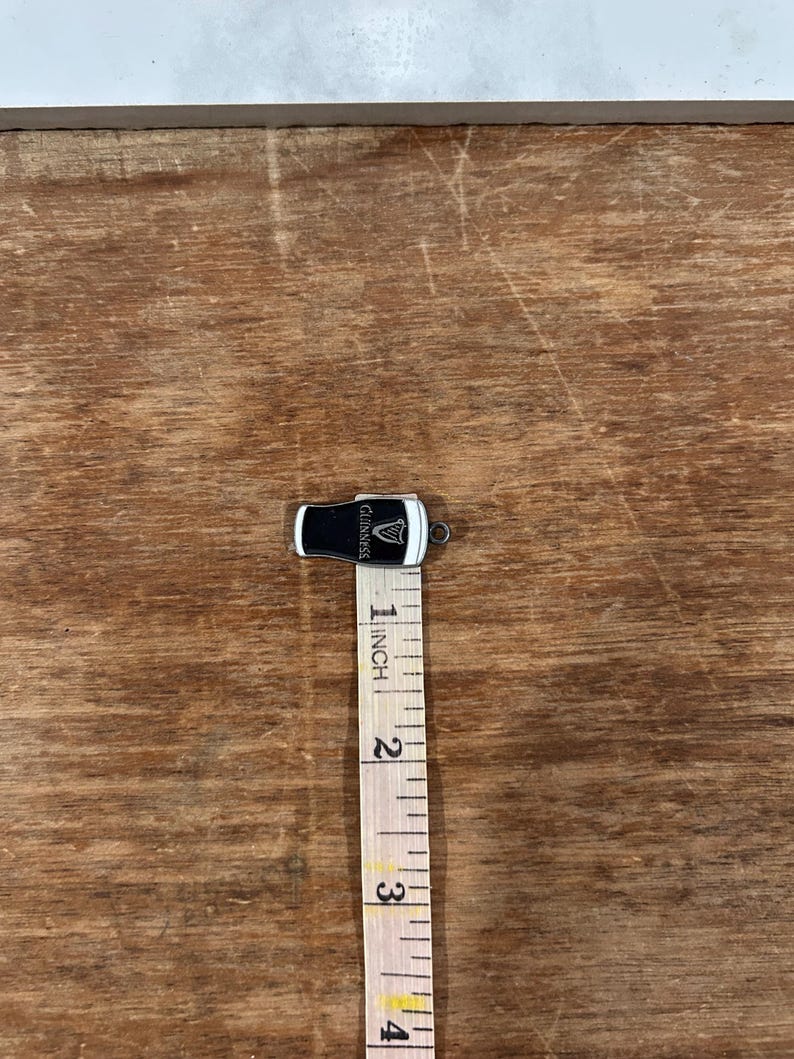 May include: A small, black and white Guinness pint glass charm with the Guinness logo. The charm is approximately 1 inch in length. The charm is on a wooden surface with a measuring tape.