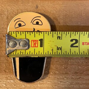 Puede incluir: Un pin de esmalte amarillo y negro de una cara de dibujos animados con un bigote negro. El pin tiene un di&aacute;metro de 2,5 cm y se mide con una cinta m&eacute;trica amarilla.