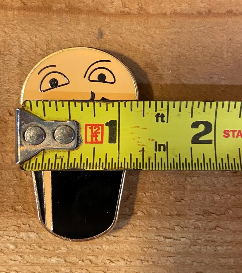 Puede incluir: Un pin de esmalte amarillo y negro de una cara de dibujos animados con un bigote negro. El pin tiene un di&aacute;metro de 2,5 cm y se mide con una cinta m&eacute;trica amarilla.