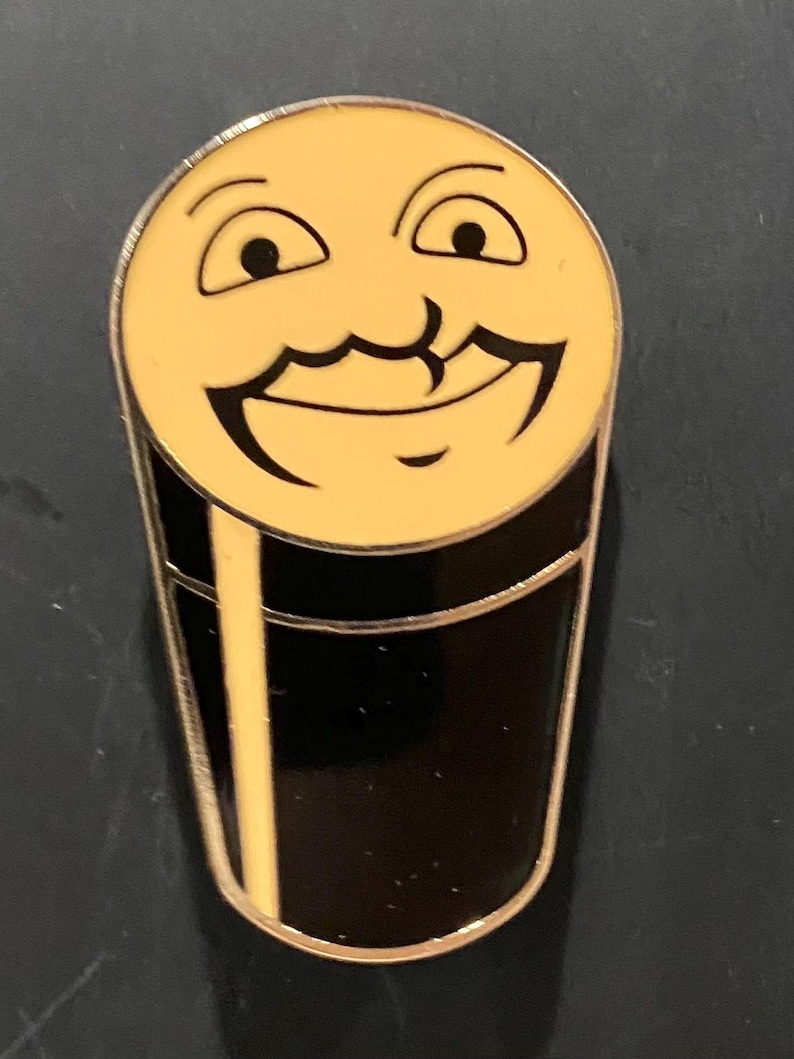 Puede incluir: Un pin de esmalte amarillo con forma de vaso de cerveza con una cara sonriente y un bigote.