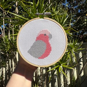 May include: A cross-stitch embroidery hoop with a pink and gray bird design. The hoop is made of wood and has a white background.