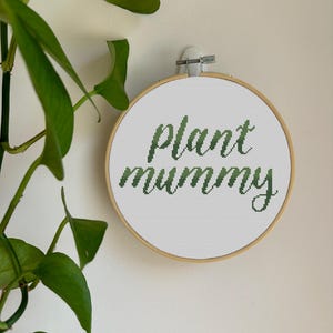 May include: A cross-stitch embroidery hoop with the words "plant mummy" in green script. The hoop is made of wood and is mounted on a white wall. Green leafy plant in the background.