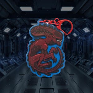 May include: Red and blue alien creature keychain with a red metal ring. The alien is in a curled up position with its tail wrapped around its body.
